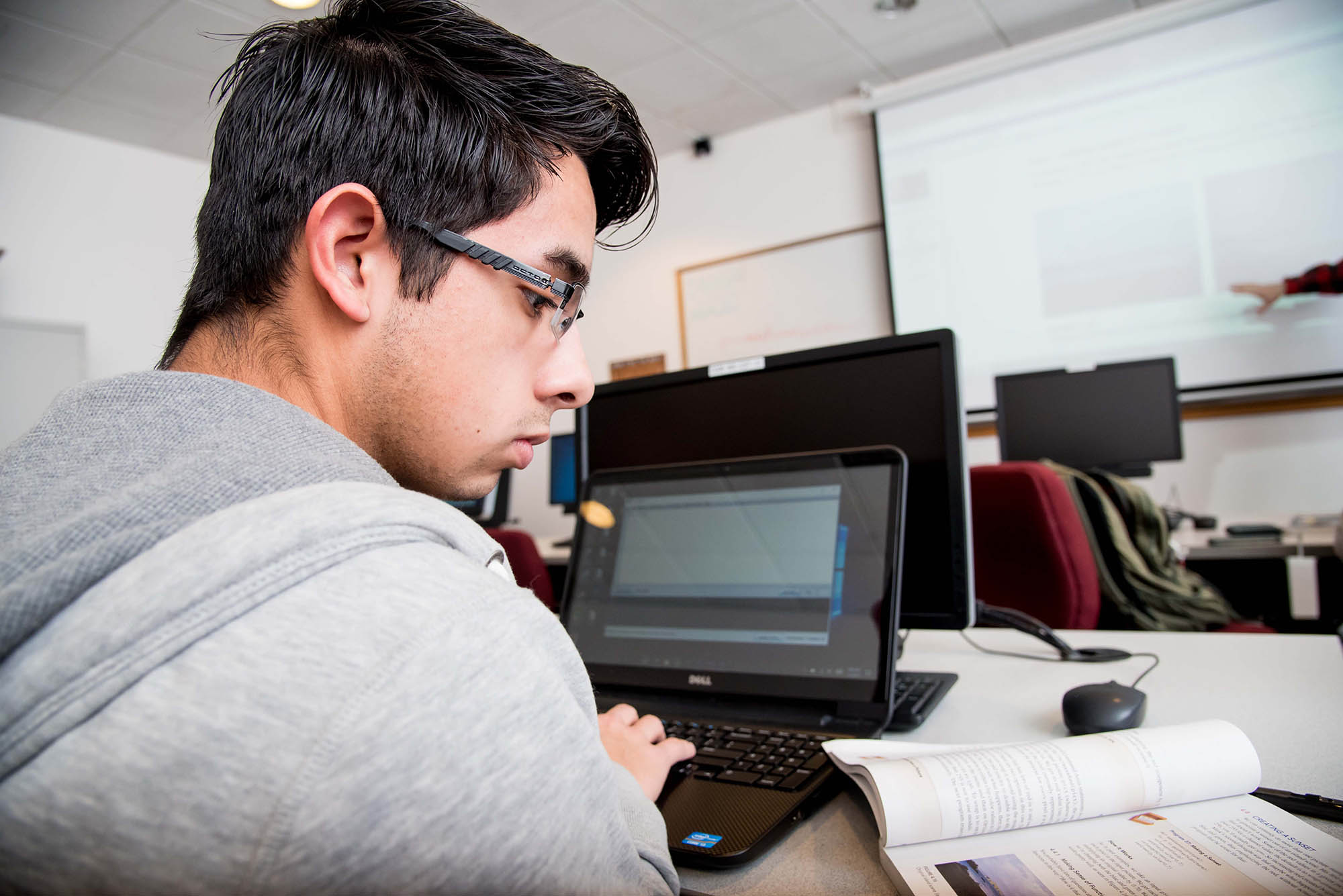 male student reading from textbook and typing on laptop